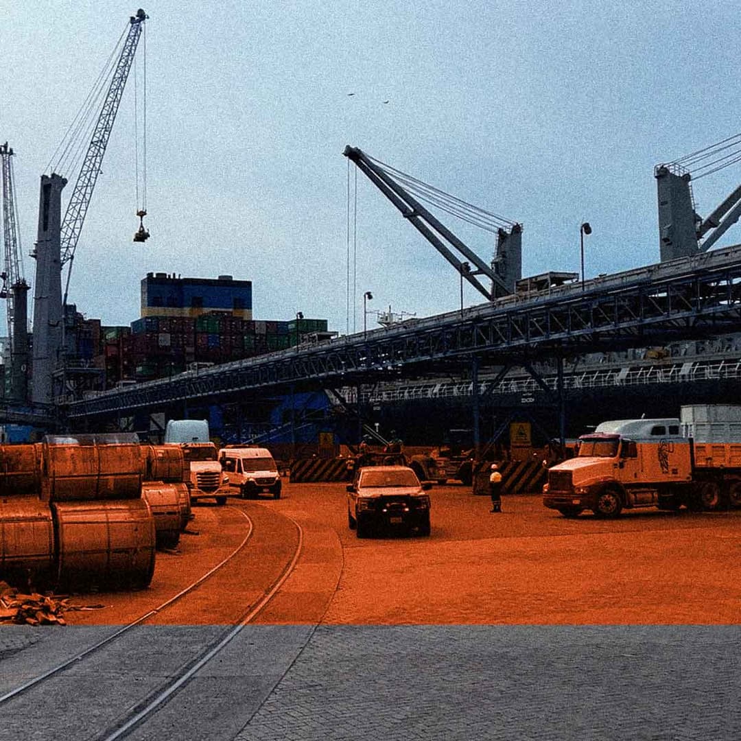 Escena de un puerto de carga con grúas, contenedores apilados, camiones y trabajadores. En primer plano se ven grandes bobinas y maquinaria de construcción. La parte inferior de la imagen está teñida de un tono anaranjado.