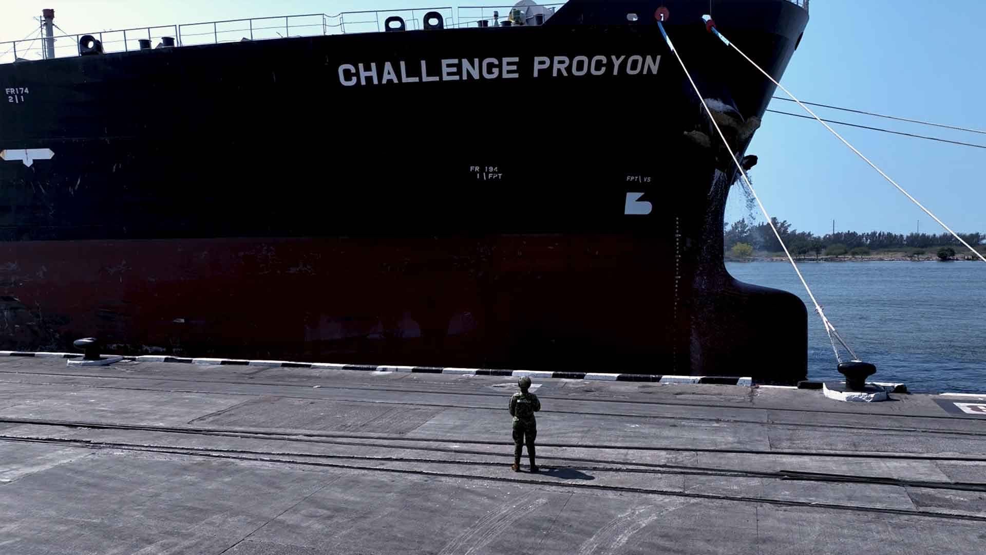 Una persona vestida de uniforme se encuentra de pie en un muelle, frente al gran buque de carga negro CHALLENGE PROCYON, amarrado en el muelle. El tiempo tranquilo y despejado contrasta con los rumores sobre el huachicol fiscal y los funcionarios cómplices que rodean el puerto.