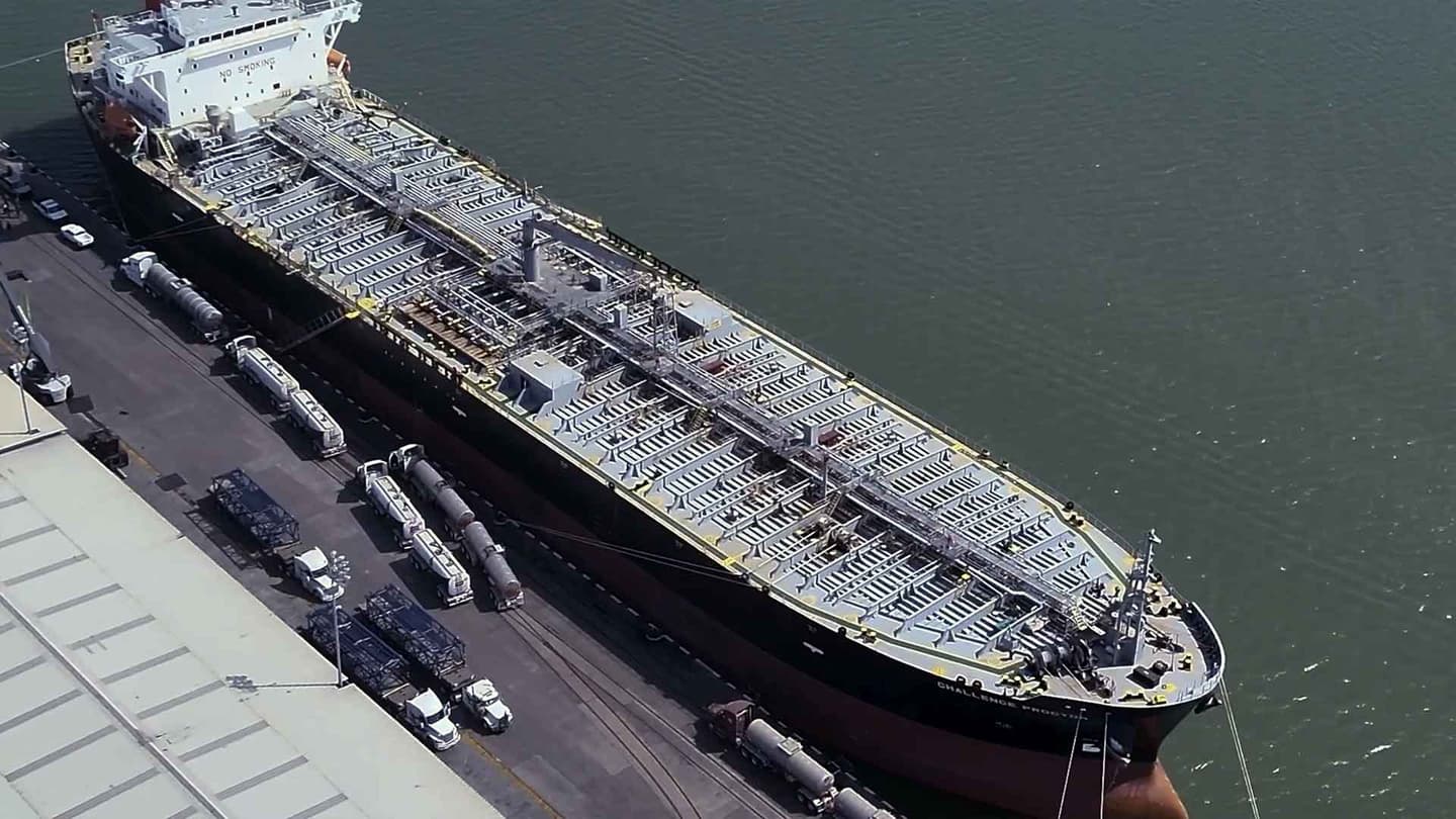 Vista aérea de un gran buque atracado en un puerto, con varios camiones y remolques aparcados junto al muelle, cerca de las instalaciones portuarias industriales.