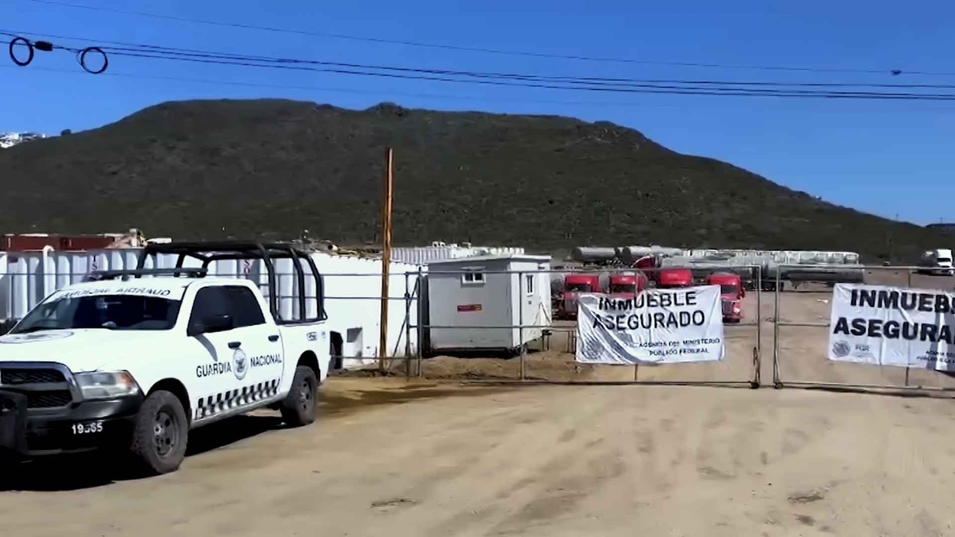 Predio privado asegurado en Ensenada. Cercado con una reja metálica en la que cuelga una pancarta con el letrero "Inmueble Asegurado", posiblemente relacionado con huachicol fiscal. Junto a la entrada hay una furgoneta blanca con la inscripción "Guardia Nacional". Se ven contenedores y pipas, con unas colinas al fondo.