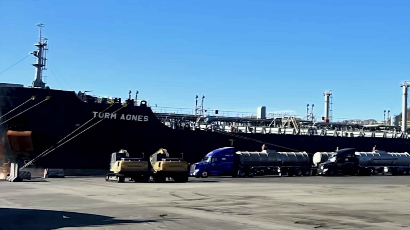 Un gran buque de carga negro llamado Torm Agnes está atracado en un puerto, donde hay varios camiones tipo pipa y vehículos de obra aparcados en las inmediaciones.