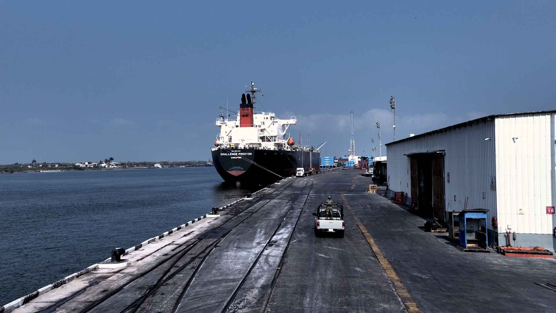 Un gran buque de carga está atracado en un puerto junto a un almacén. El muelle se adentra en el agua y hay vehículos aparcados a lo largo de él, mientras que, al fondo, bajo un cielo azul claro, se apilan contenedores de transporte.
